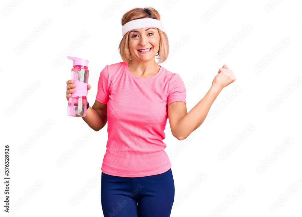 Young blonde woman wearing sportswear holding water bottle screaming proud, celebrating victory and success very excited with raised arms Young blonde woman wearing sportswear holding water bottle screaming proud, celebrating victory and success very excited with raised arms