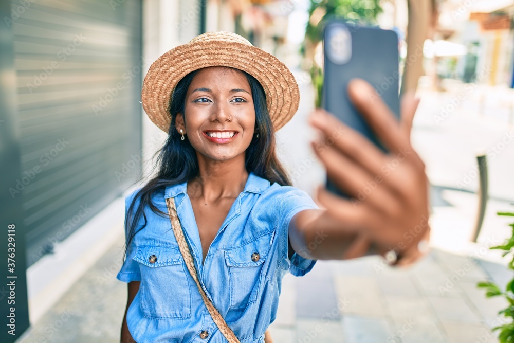 Young beautiful indian woman wearing summer hat smiling happy making selfie by the smartphone at the city.
