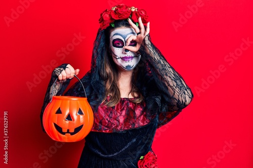 Young woman wearing day of the dead costume holding pumpkin smiling happy doing ok sign with hand on eye looking through fingers