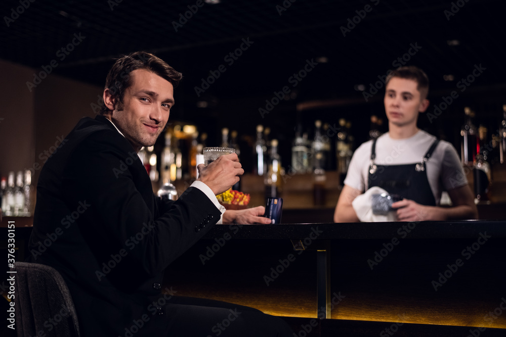 a handsome man poses at the bar with a cocktail in his hands against the background of the bartender and bottles.