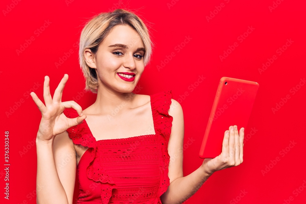 Young beautiful blonde woman holding touchpad doing ok sign with fingers, smiling friendly gesturing excellent symbol Young beautiful blonde woman holding touchpad doing ok sign with fingers, smiling friendly gesturing excellent symbol