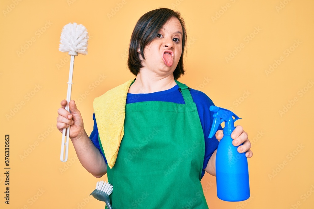 Brunette woman with down syndrome wearing apron holding scourer and toilet brush sticking tongue out happy with funny expression.