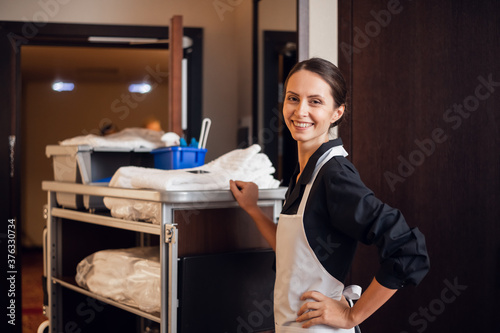 A smiling hotel maid with cleaning cart and cleaning supplies, looking at the camera and posing. photo