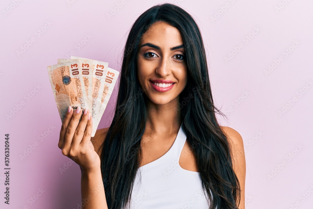 Beautiful hispanic woman holding united kingdom 10 pounds looking positive and happy standing and smiling with a confident smile showing teeth Beautiful hispanic woman holding united kingdom 10 pounds looking positive and happy standing and smiling with a confident smile showing teeth