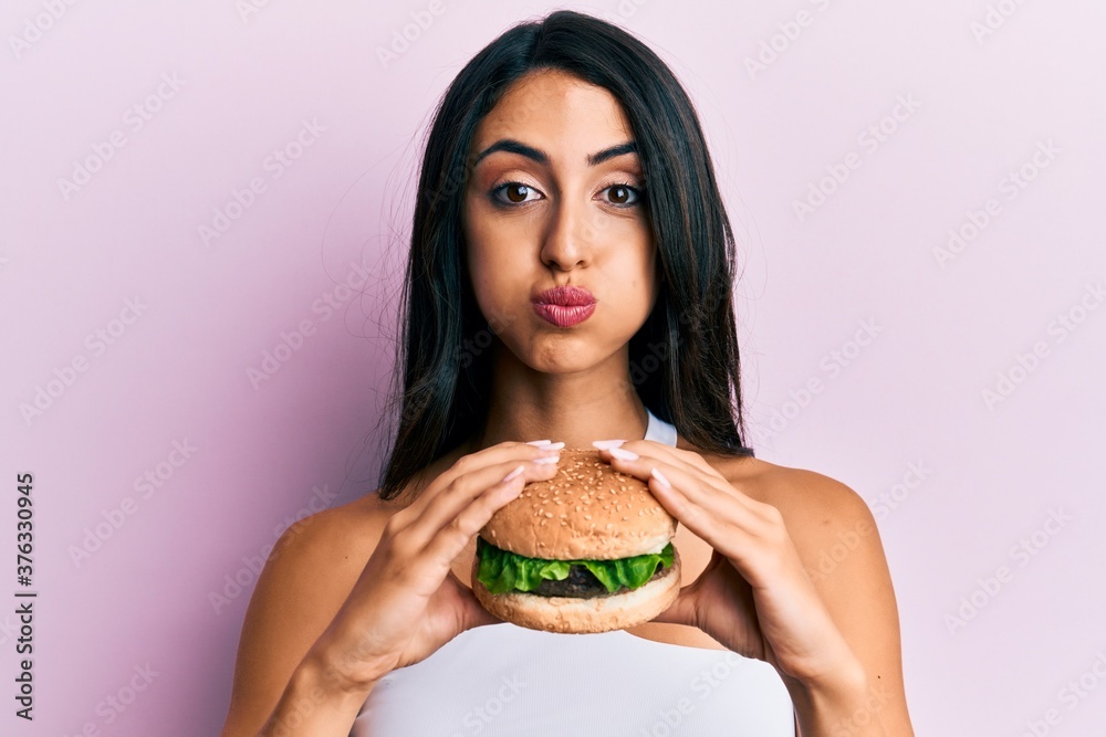 Beautiful hispanic woman eating a tasty classic burger puffing cheeks with funny face. mouth inflated with air, catching air.