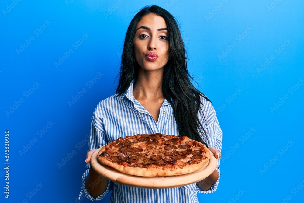 Beautiful hispanic woman holding tasty pepperoni pizza looking at the camera blowing a kiss being lovely and sexy. love expression. Beautiful hispanic woman holding tasty pepperoni pizza looking at the camera blowing a kiss being lovely and sexy. love expression.