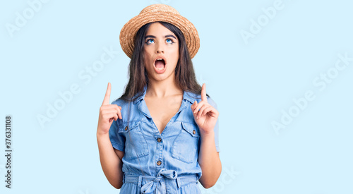 Brunette teenager girl wearing summer hat amazed and surprised looking up and pointing with fingers and raised arms. © Krakenimages.com