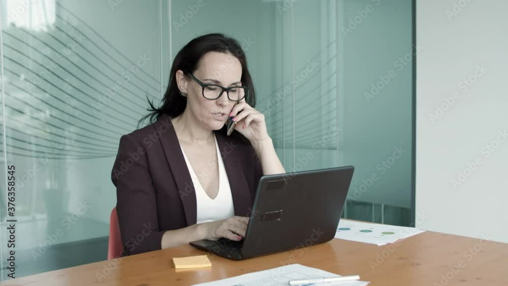 Busy female executive wearing glasses, using laptop in office while talking on cellphone, speaking, typing, looking at screen. Medium shot. Communication or multitasking concept Busy female executive wearing glasses, using laptop in office while talking on cellphone, speaking, typing, looking at screen. Medium shot. Communication or multitasking concept
