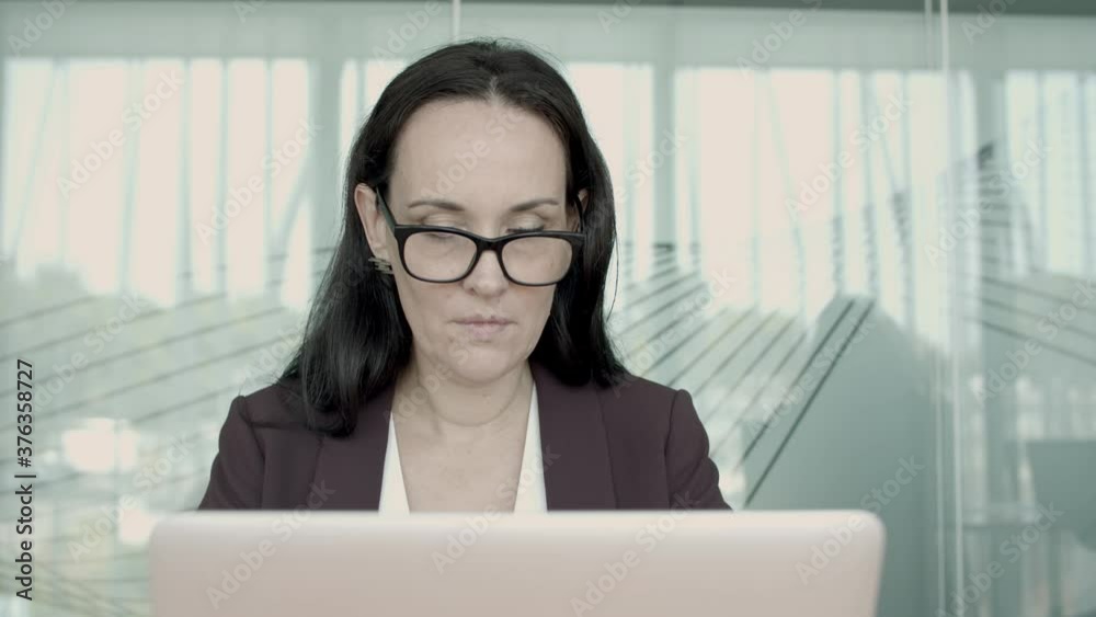 Focused busy female professional wearing suit and glasses, typing on laptop in office, using computer at workplace. Front view. Communication or internet technology concept