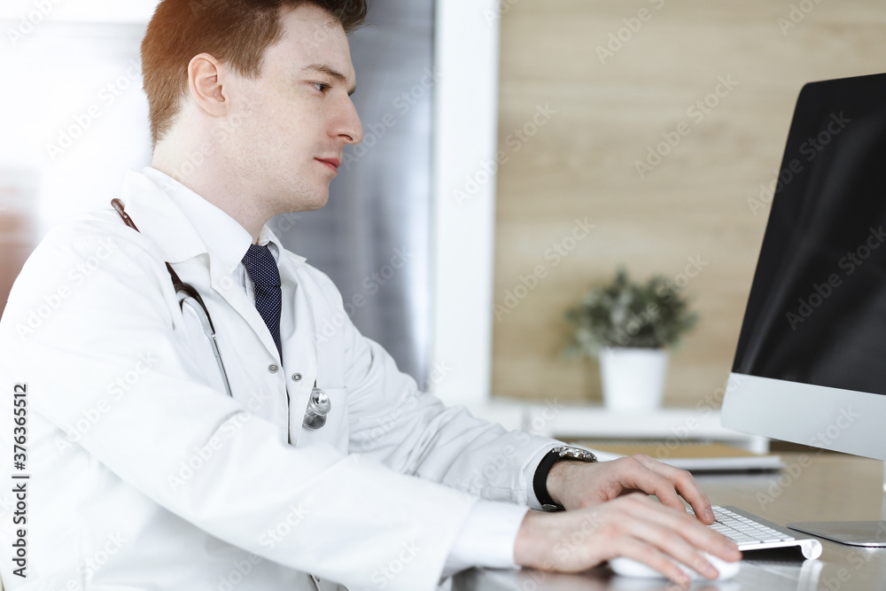 Doctor man sitting behind a computer at workplace in sunny clinic