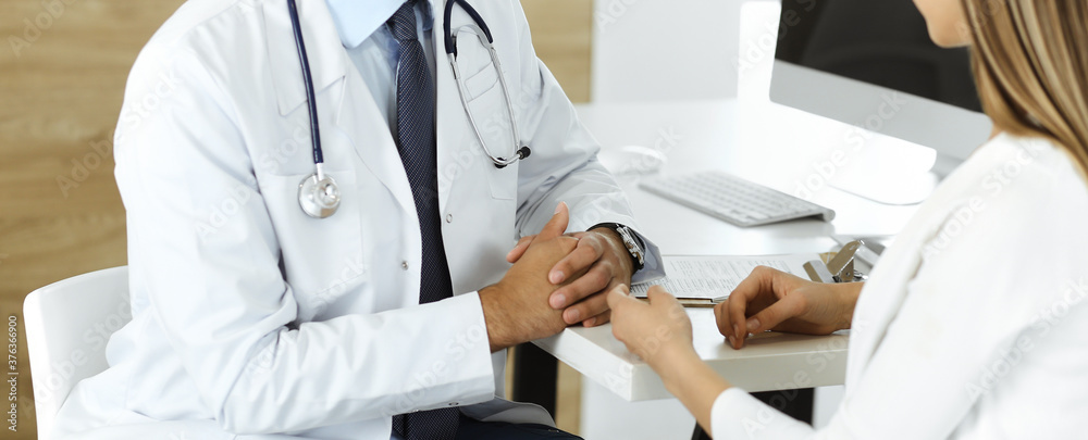 Unknown doctor and patient discussing something while sitting at the desk in clinic, close-up. Concept of medicine and comfortable service in clinic Unknown doctor and patient discussing something while sitting at the desk in clinic, close-up. Concept of medicine and comfortable service in clinic