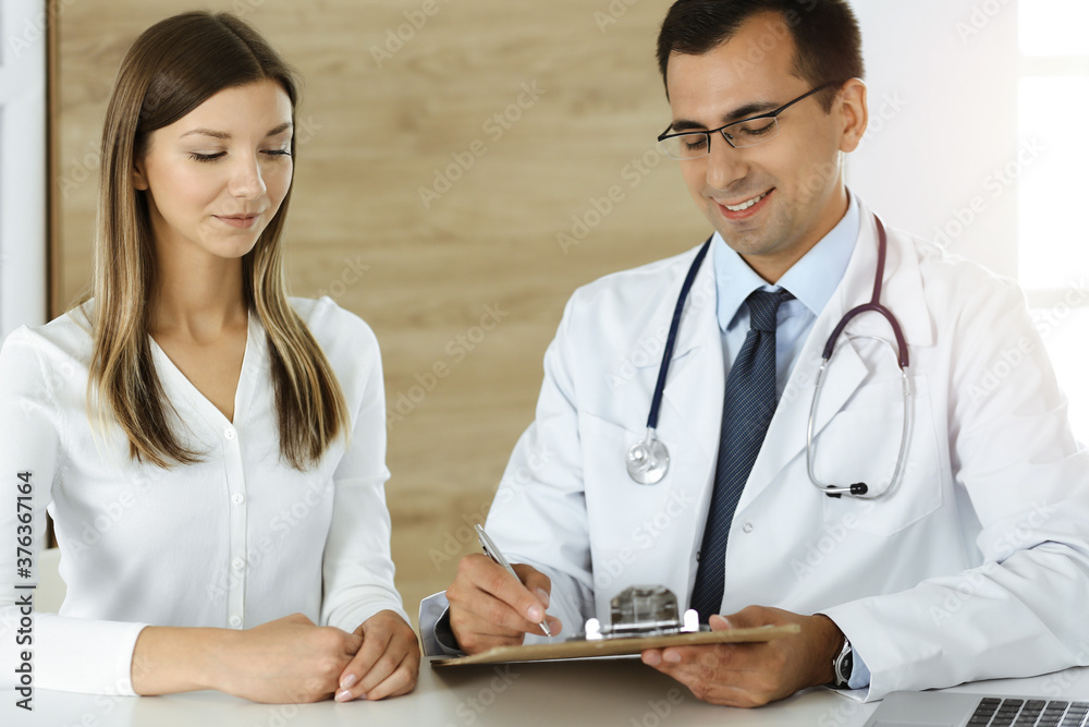 Doctor and patient discussing medical exam results while sitting at the desk in sunny clinic Doctor and patient discussing medical exam results while sitting at the desk in sunny clinic
