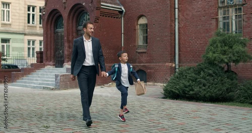 Little boy with bag jumping hapilly while going and holding hand in hand his smiling father. Cheerful man in suit taking his son to school. Concept of children and education. photo