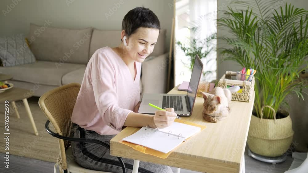 happy short-haired woman with bluetooth earpiece writes in paper notebook sitting at table with modern laptop and cute cat in light living room happy short-haired woman with bluetooth earpiece writes in paper notebook sitting at table with modern laptop and cute cat in light living room