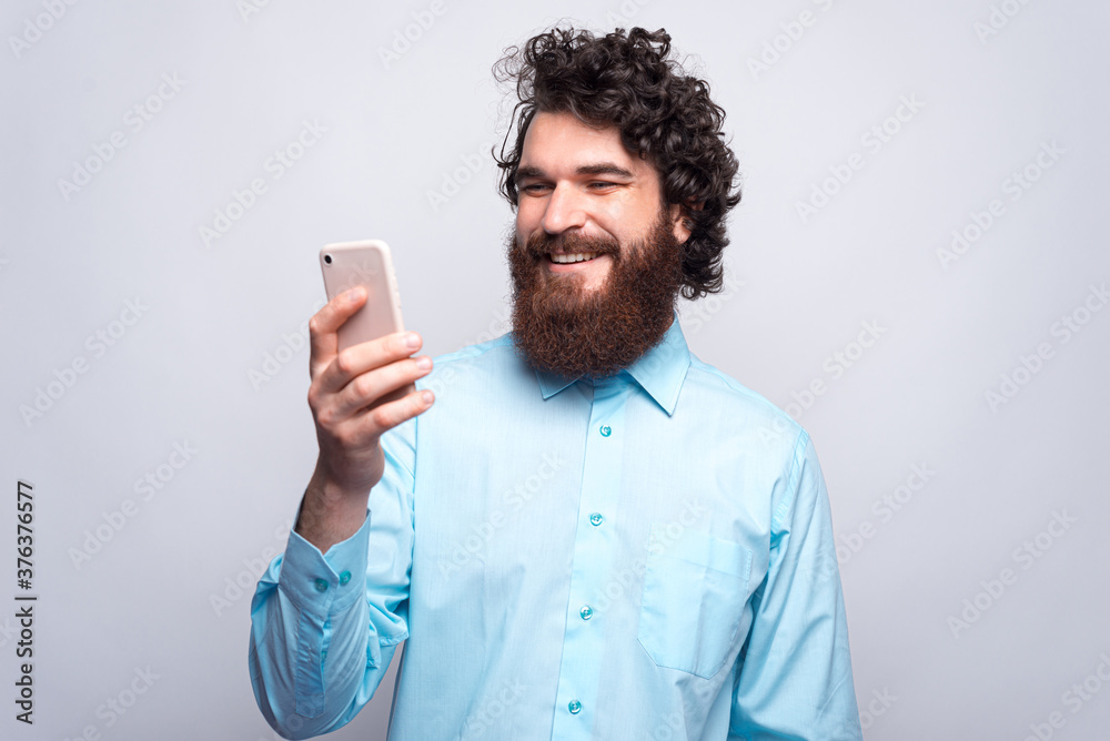 Cheerful young bearded hipster man in blue shirt using his smartphone and smiling Cheerful young bearded hipster man in blue shirt using his smartphone and smiling