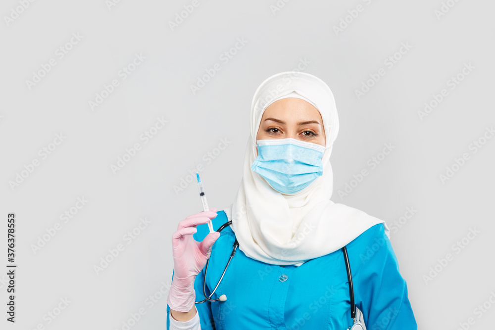 Muslim doctor or nurse in hijab and medical face mask holding a syringe on a gray background. the concept of vaccination of the population or injection plastic of the face