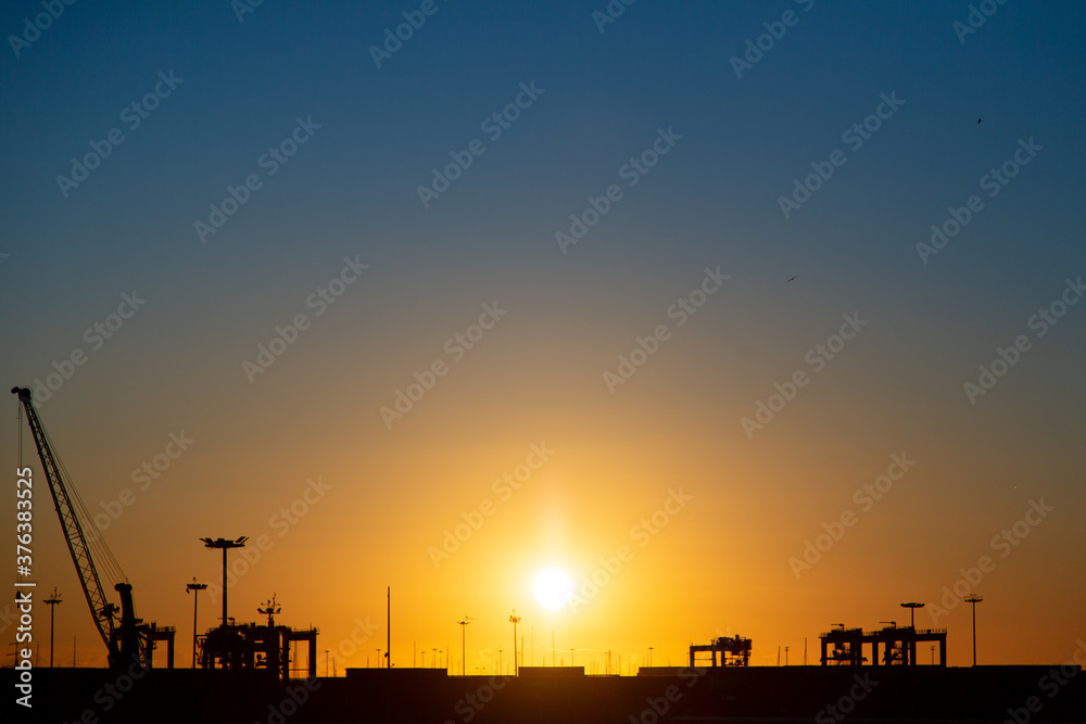 Sunset behind the sea and over a seaport with ships and port machinery. Valencia, Spain. With orange and blue colors