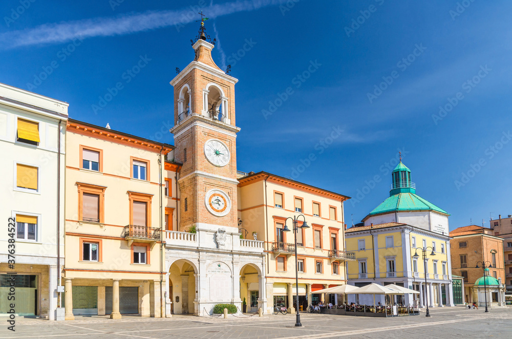 Piazza Tre Martiri Three Martyrs square with traditional buildings with clock and bell tower in old historical touristic city centre Rimini with blue sky background, Emilia-Romagna, Italy