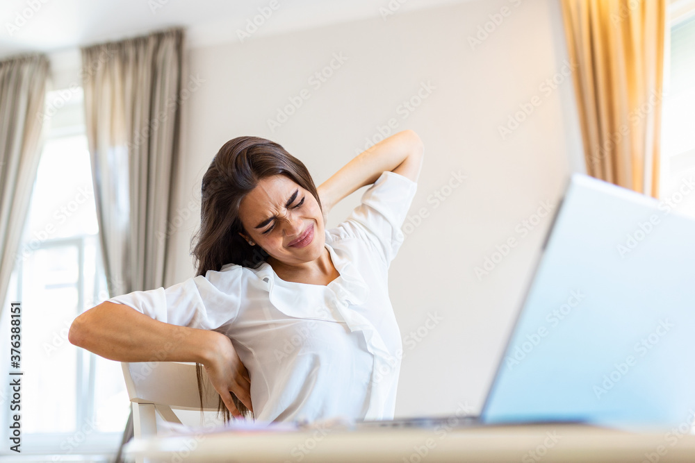 Portrait of young stressed woman sitting at home office desk in front of laptop, touching aching back with pained expression, suffering from back, spine pain after working on pc Portrait of young stressed woman sitting at home office desk in front of laptop, touching aching back with pained expression, suffering from back, spine pain after working on pc