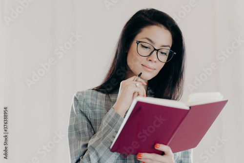 Serious brunette female management consultant uses notepad for planning organization, holds pen and writes down notes or list to do, wears formal clothing, has serious expression, looks in notebook