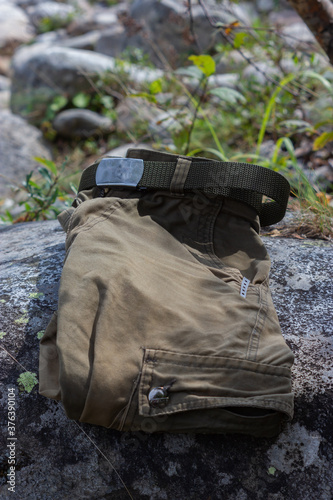 Folded trousers in military style with a belt with a shiny buckle, with a knife in a pocket lying on a stone