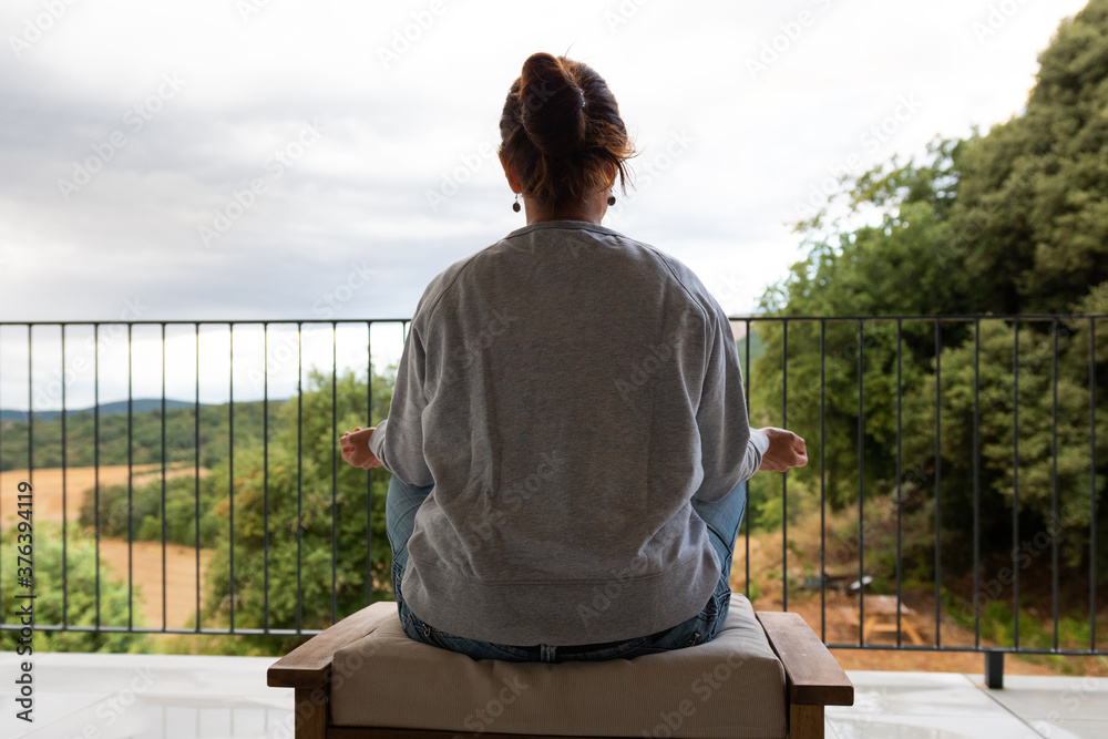 Mujer sentada meditando en el balcón del hogar con las piernas recogidas y los brazos extendidos
