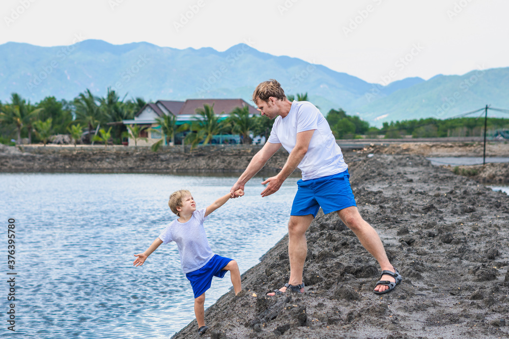 Dad helps son go down to lake water, explains safety rules. Family together walk play. Home natural child education, fathers day, fatherly responsibilities, influence on formation of son worldview Dad helps son go down to lake water, explains safety rules. Family together walk play. Home natural child education, fathers day, fatherly responsibilities, influence on formation of son worldview