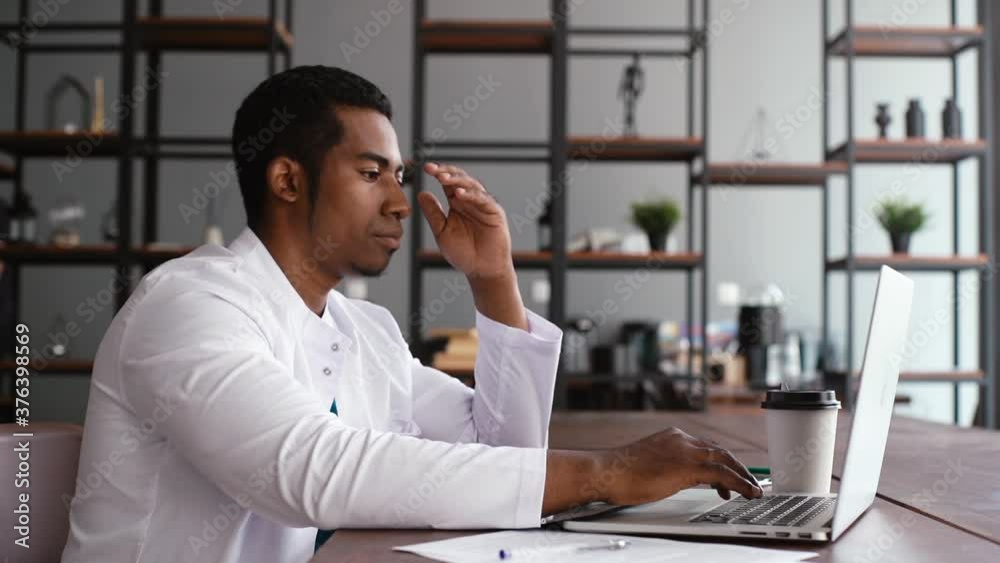 Side view of focused African American black male doctor in white gown having head pain while working typing on laptop computer, sitting at the desk on workplace in light office room at hospital. Side view of focused African American black male doctor in white gown having head pain while working typing on laptop computer, sitting at the desk on workplace in light office room at hospital.