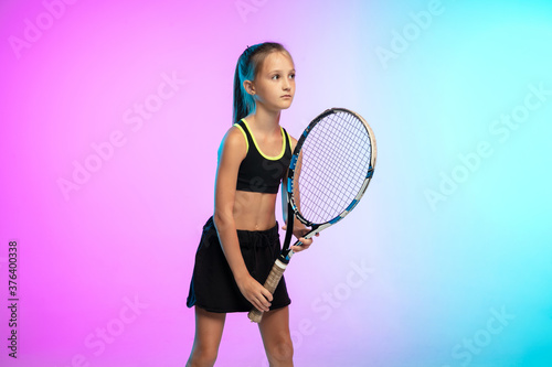 Winner. Little tennis girl in black sportwear isolated on gradient background in neon light. Little caucasian model, sport kid training in motion and action. Sport, movement, childhood concept.