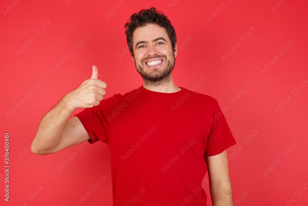 Young handsome caucasian man wearing t-shirt over isolated red background doing happy thumbs up gesture with hand. Approving expression looking at the camera showing success. Young handsome caucasian man wearing t-shirt over isolated red background doing happy thumbs up gesture with hand. Approving expression looking at the camera showing success.