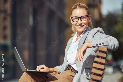 Beautiful succesful middle aged business woman wearing eyeglases sitting on the bench outdoors and working online on laptop photo