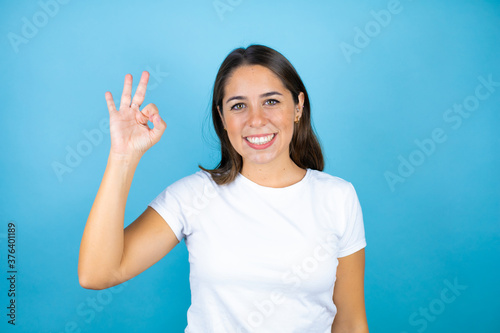 Young beautiful woman over isolated blue background doing ok sign with fingers and smiling, excellent symbol