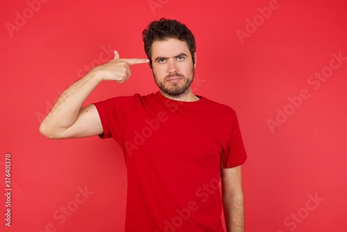 Unhappy  Young handsome caucasian man wearing t-shirt over isolated red wall makes suicide gesture and imitates gun with hand, curves lips keeps two fingers on temple, shoots, being tired 