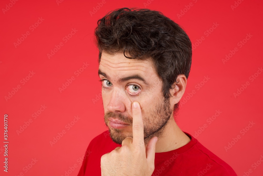 Young handsome caucasian man wearing t-shirt over isolated red background Pointing to the eye watching you gesture, suspicious expression.
