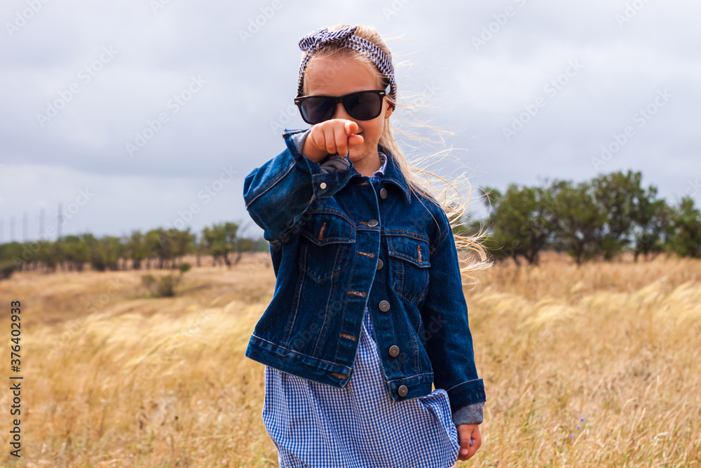 Adorable little girl in denim jacket, black sunglasses, blue plaid dress in yellow grass field. Happy stylish long blonde hair child on countryside landscape. Cute kid walking outdoor rural road trip.