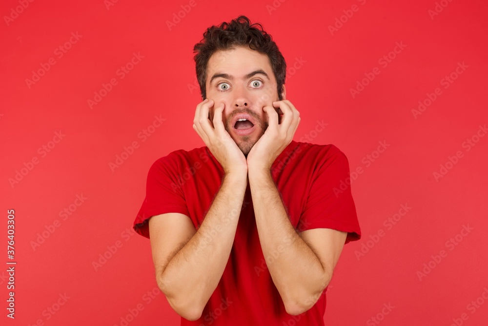 Stupefied Young handsome caucasian man wearing t-shirt over isolated red background, expresses excitement and thrill, keeps jaw dropped, hands on cheeks, has eyes popped out