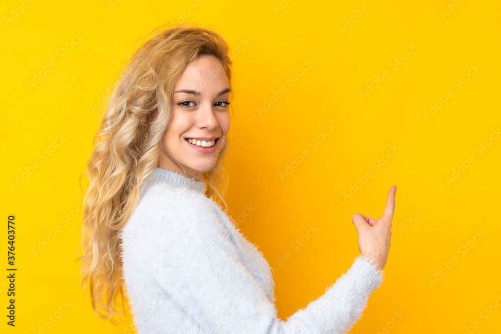 Young blonde woman isolated on yellow background pointing back Young blonde woman isolated on yellow background pointing back