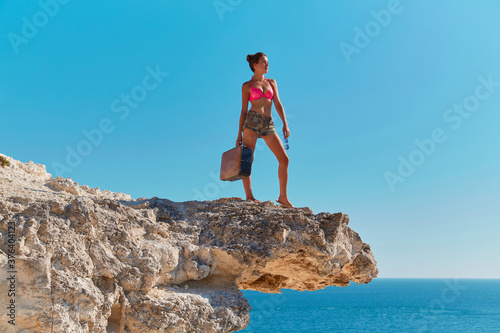 Woman in beachwear with suitcase and glasses. Ocean view from cliff. Time for travel or Summer holiday. Travel desire.