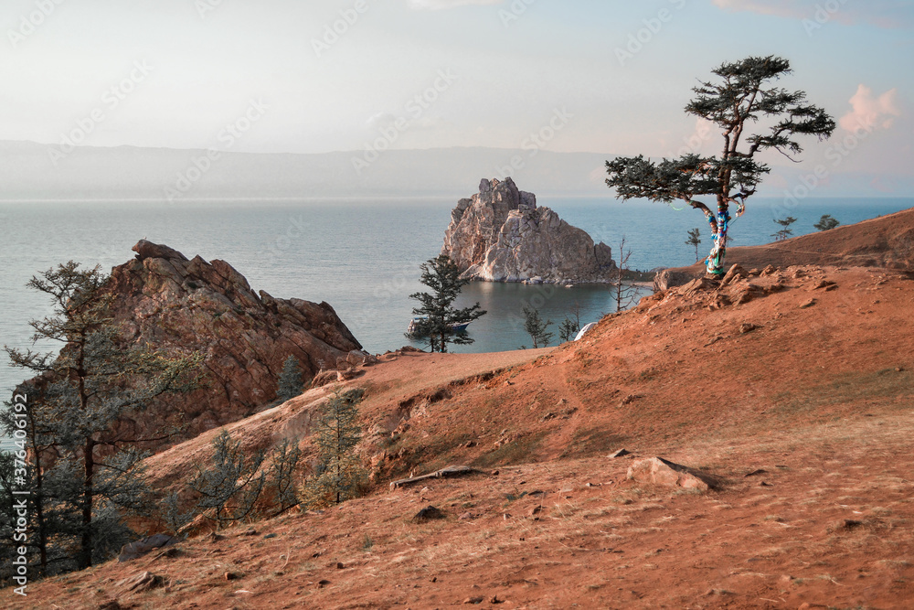 Cape Shamanka rock in Lake Baikal among the grassy steppes with coniferous larch trees, against the background of mountains in sunset llight