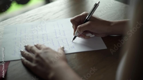 Young woman writes love letter on white paper by pen to her beloved groom, sits at the table in room near balcony. Green grass outside the balcony. Wedding preparations. Bride’s vow. Slow motion.