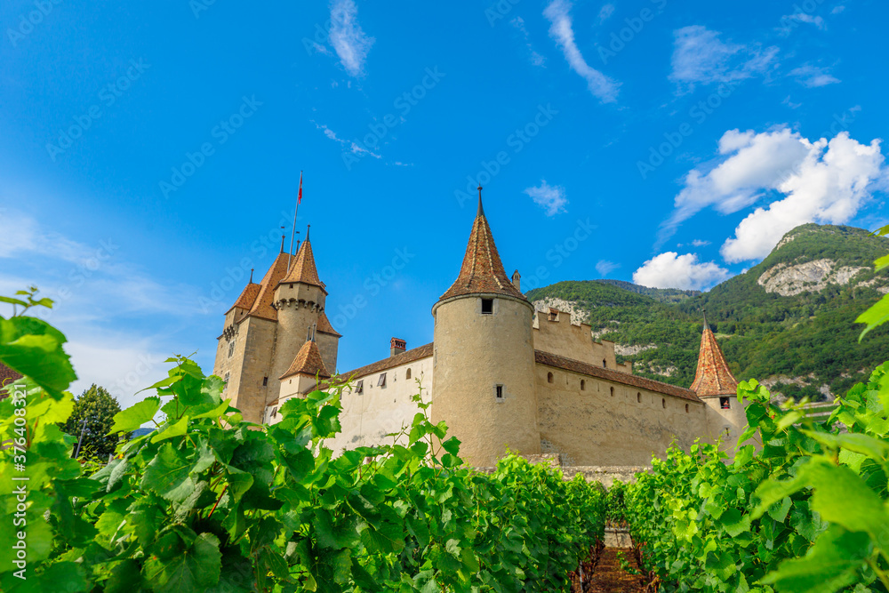 Fototapeta premium Grape leaves in foreground and rows of vines surround Chateau d'Aigle in Canton Vaud, Switzerland. Aigle Castle overlooking terraced vineyards and Swiss Alps. Wine region with popular tasting tours.