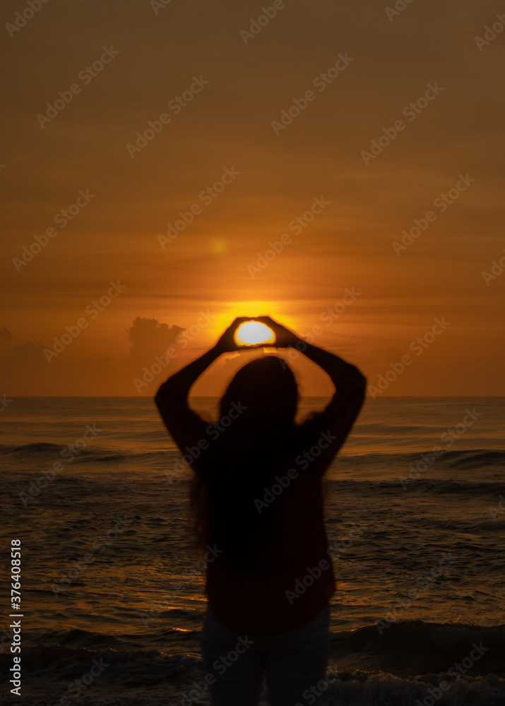 a woman poses as a heart shape by keeping the sun between her hands on the seashore.