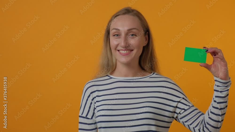 A beautiful young woman is holding a blank green card in her hands, looking at the camera and smiling. Yellow bright background. 4K video. A beautiful young woman is holding a blank green card in her hands, looking at the camera and smiling. Yellow bright background. 4K video.
