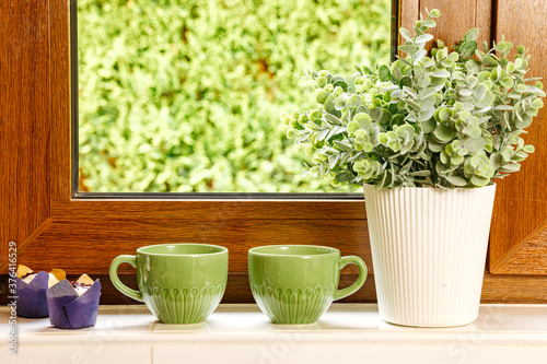 Two green cups with tea stand on a white windowsill next to a plant.