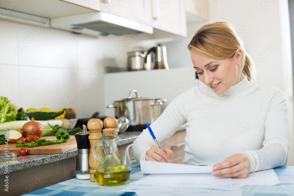 Happy woman sitting with financial papers in kitchen and smiling