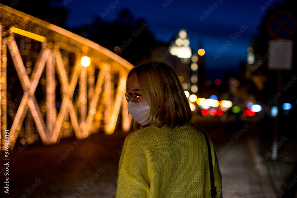 A young woman in a mask standing in the city at night. Coronavirus concept. Woman wearing medical mask in the city. Bokeh background. Health issues. Covid 19. Night life with coronavirus.