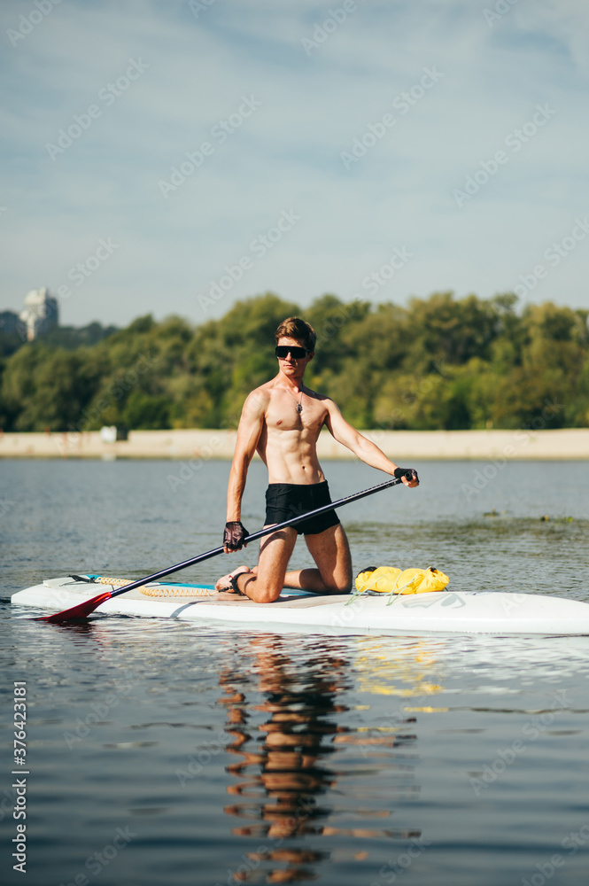 Muscular athletic young man spends free time in summer on the river, paddles on the sup board. Workout on sup board on a sunny day. Vertical