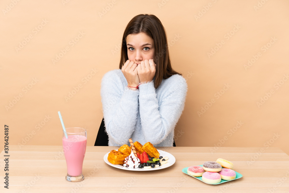 Teenager girl eating waffles isolated on beige background nervous and scared putting hands to mouth Teenager girl eating waffles isolated on beige background nervous and scared putting hands to mouth