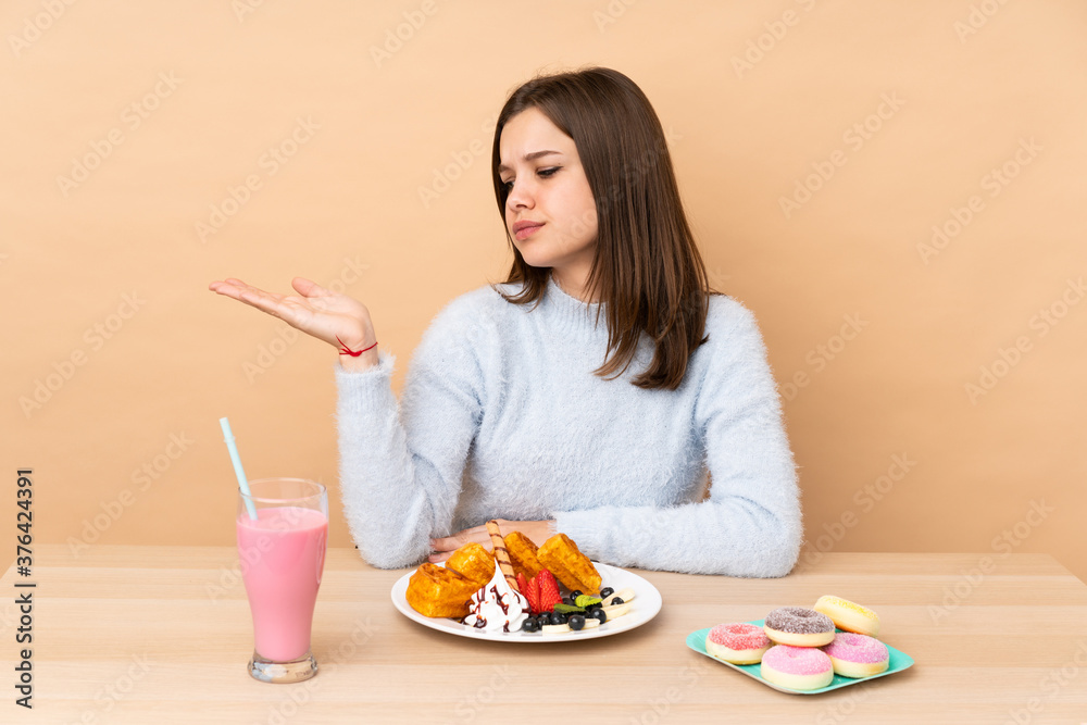 Teenager girl eating waffles isolated on beige background holding copyspace with doubts Teenager girl eating waffles isolated on beige background holding copyspace with doubts