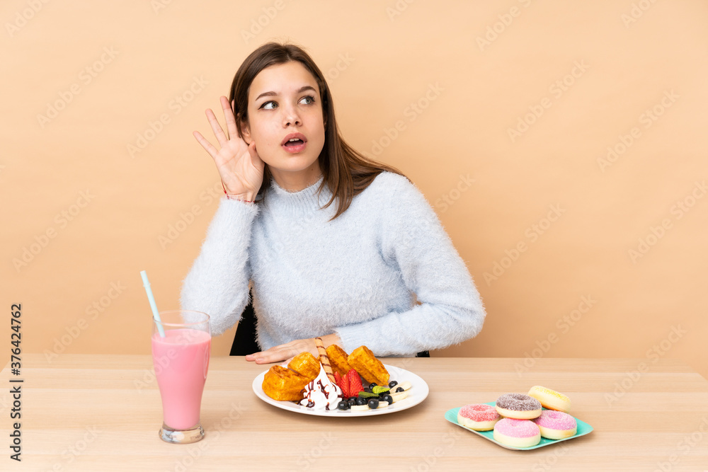 Teenager girl eating waffles isolated on beige background listening to something by putting hand on the ear